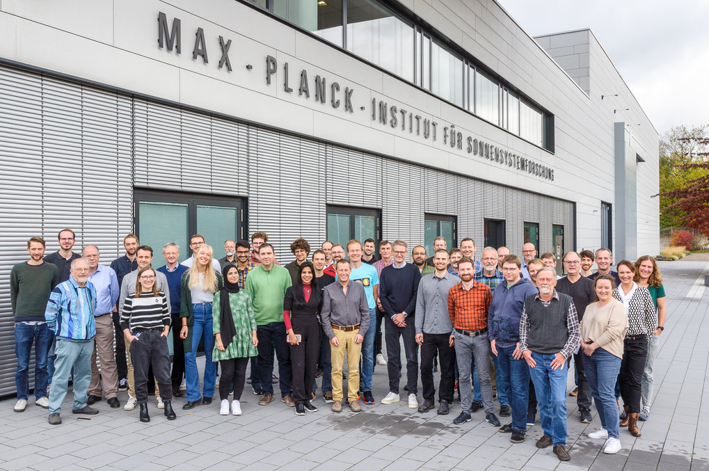 A group of about fifty people of various ages and genders, many of them appearing to be of european origin, staning in front of a gray building front and looking at the camera.